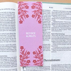 Può includere: Un segnalibro rosa con un motivo floreale e le parole "REJOICE ALWAYS" in bianco, posto in un libro. Il segnalibro ha un motivo floreale decorativo in rosso. Le pagine del libro sono visibili.