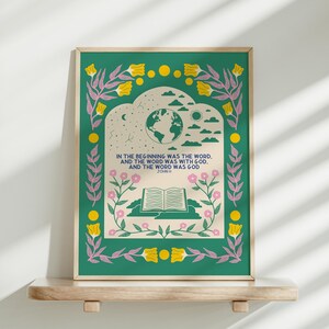 May include: A framed art print with a green border and yellow and pink floral details. The artwork displays the text "IN THE BEGINNING WAS THE WORD, AND THE WORD WAS WITH GOD, AND THE WORD WAS GOD" and an illustration of the Earth, clouds, and an open book.