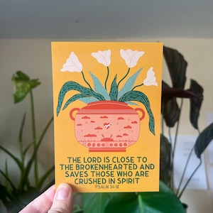 May include: A yellow card with a white flower illustration in a pink pot with a heart and clouds. The text reads "The Lord is close to the brokenhearted and saves those who are crushed in spirit. Psalm 34:18"