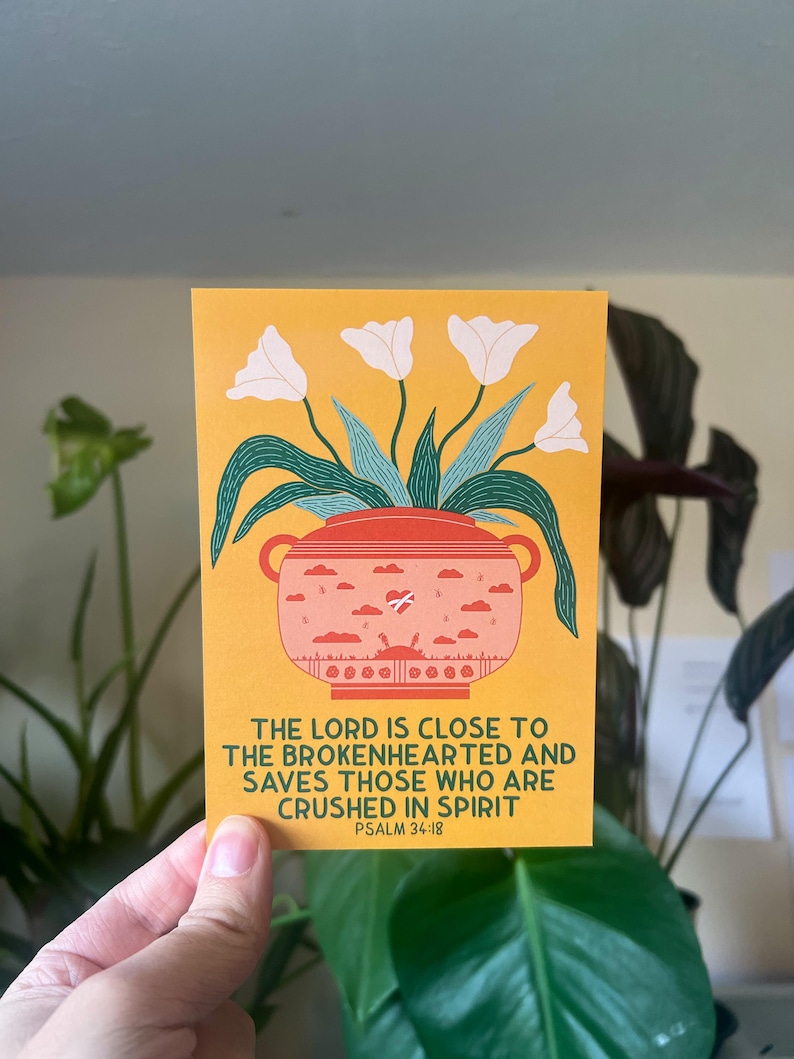 May include: A yellow card with a white flower illustration in a pink pot with a heart and clouds. The text reads "The Lord is close to the brokenhearted and saves those who are crushed in spirit. Psalm 34:18"
