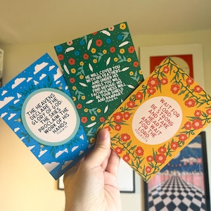May include: Three colorful cards with inspirational quotes from the Bible. The cards feature floral designs and are printed on a white background. The text on the cards reads: "The heavens declare the glory of God: the skies proclaim the work of his hands." (Psalm 19:1), "He will cover you with his feathers, and under his wings you will find refuge: his faithfulness will be your shield and rampart." (Psalm 91:4), and "Wait for the Lord: be strong and take heart and wait for the Lord." (Psalm 27:14)