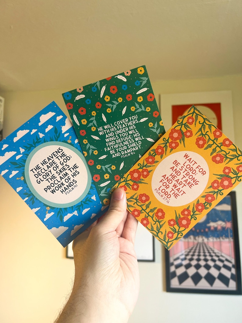 May include: Three colorful cards with inspirational quotes from the Bible. The cards feature floral designs and are printed on a white background. The text on the cards reads: "The heavens declare the glory of God: the skies proclaim the work of his hands." (Psalm 19:1), "He will cover you with his feathers, and under his wings you will find refuge: his faithfulness will be your shield and rampart." (Psalm 91:4), and "Wait for the Lord: be strong and take heart and wait for the Lord." (Psalm 27:14)
