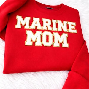 Pode incluir: Um moletom vermelho com as palavras "MARINE MOM" em letras grandes e brancas com bordas douradas. O moletom é feito de um material macio e está dobrado, com as mangas visíveis. O fundo é um tecido branco macio.