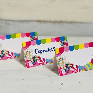 May include: Four colorful candy-themed tent cards with a white background. The cards feature a vibrant design of lollipops, gumball machines, and cupcakes. One card has the word "Cupcakes" written on it in blue.