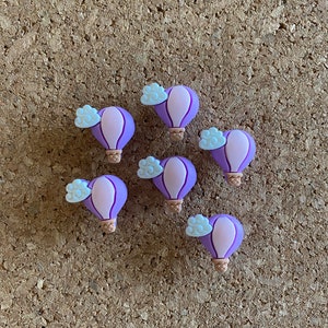 May include: Eight purple hot air balloons with white clouds, each with a brown basket, on a corkboard background.