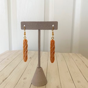 May include: A pair of novelty earrings shaped like miniature baguettes. The earrings are a light brown colour with a textured surface, and hang from gold-coloured hooks. The earrings are displayed on a grey jewellery stand against a light wood background.