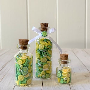 May include: Three clear glass bottles with cork stoppers filled with yellow, green, and white decorative pieces. The bottles contain lemon and lime slices, and small white beads. The tallest bottle has a white ribbon and a lemon slice charm.