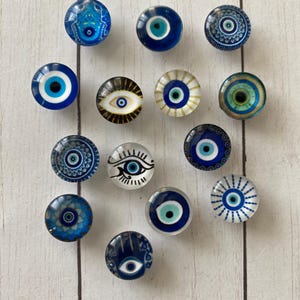 May include: A collection of round, glass-like decorative knobs featuring various designs of the evil eye in shades of blue, white, and gold. The knobs are arranged on a light wood-grain surface.