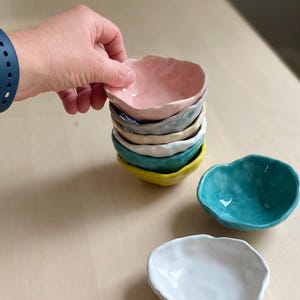 May include: A collection of small, irregularly shaped ceramic bowls in various colours, including pink, blue, yellow, and white. One hand is holding the top bowl. The bowls have a textured, organic design.