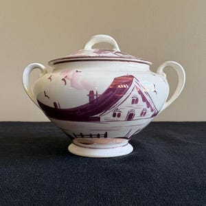 May include: A white ceramic sugar bowl with a lid, featuring a purple illustration of a house and landscape. The bowl has two handles and a matching lid with a small handle. The design includes birds and a fence.