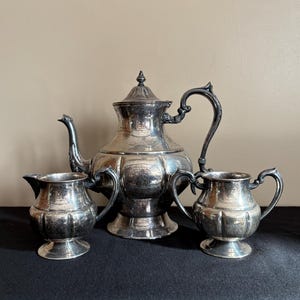 May include: A silver tea set, including a teapot, creamer, and sugar bowl, is displayed on a black surface. The teapot has a curved spout and ornate handle. The creamer and sugar bowl have matching handles.