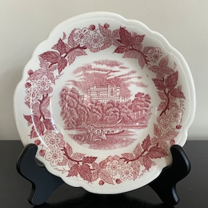 May include: A white ceramic plate with a pink floral design and a pink image of a castle in the center. The plate has a scalloped edge.