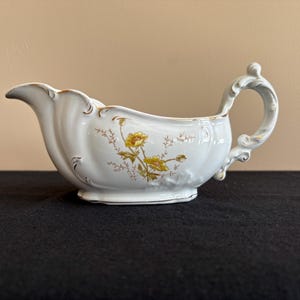 May include: White porcelain gravy boat with a scalloped rim and ornate handle. The boat features a floral design in yellow and brown, with gold trim accents. A classic piece for serving sauces and gravies.