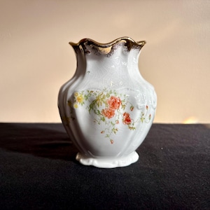 May include: White ceramic vase with a ruffled gold rim and floral design. The vase features a delicate pattern of red, yellow, and green flowers. The vase is approximately 6 inches tall.