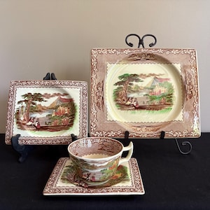 May include: A vintage tea set featuring a cup, saucer, and two square plates, all with a brown and cream landscape design. The plates are displayed on black metal stands. The cup and saucer are placed in front of the plates.