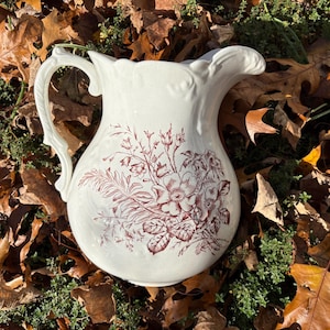May include: Cream-colored ceramic pitcher with a floral design in shades of burgundy. The pitcher has a curved handle and a scalloped rim. The floral pattern includes flowers and leaves. The pitcher is sitting on a bed of leaves.