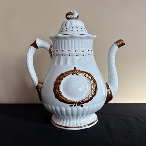May include: White ceramic teapot with gold accents. The teapot features a decorative gold wreath design on the front, a twisted handle, and a matching lid with a gold finial. The spout and handle are trimmed with gold.