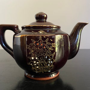 May include: A small brown teapot with gold and white floral designs. The teapot has a rounded body and a short spout.