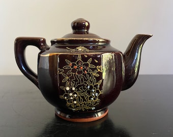 May include: A small brown teapot with gold and white floral designs. The teapot has a rounded body and a short spout.