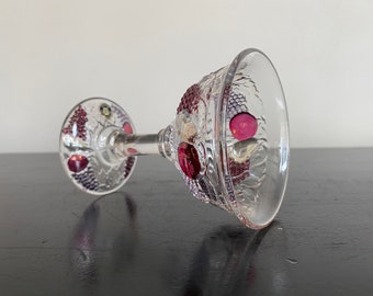Può includere: Calice in vetro trasparente con stelo e coppa decorativi. Il calice è ornato da grappoli d'uva in rilievo e gioielli colorati nelle tonalità del rosso, viola e rosa. Il calice è appoggiato su una superficie scura.