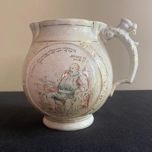 May include: A white ceramic pitcher with a dragon-shaped handle. The pitcher is decorated with a scene from the play "Henry IV, Part II" by William Shakespeare. The text on the pitcher reads "In streets, I'll fight thee, villainous company, Though in the spirit of me, I'll not be forc'd to yield."  Henry IV, Act III, Sc. 5.