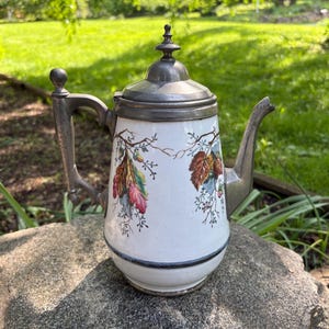 May include: Antique white porcelain coffee pot with a metal lid, handle, and spout. The pot is decorated with colorful leaf and branch designs. The pot is sitting on a gray stone.