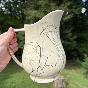 May include: Cream-colored ceramic pitcher with a curved spout and handle. The pitcher is decorated with black, abstract scribble lines. The pitcher is held by a person in front of a green background.