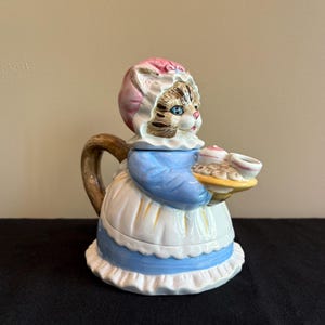 May include: A ceramic teapot in the shape of a cat wearing a bonnet and apron, holding a tray of treats. The teapot features a brown handle and spout, with blue, white, and pink accents. The cat's face is detailed with blue eyes and tabby markings.