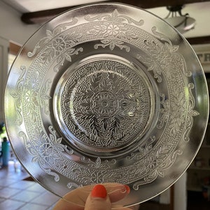 May include: A clear glass plate with a floral pattern etched into the surface. The plate has a raised center with a more intricate design.