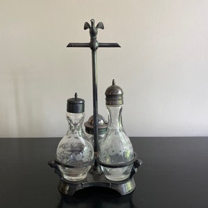 May include: Silver condiment set with three glass bottles with etched floral designs. The bottles are on a silver stand with a bird finial.