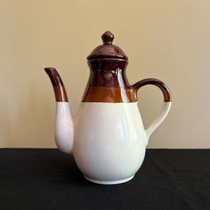 May include: A ceramic teapot featuring a glossy finish with a white base, a brown middle section, and a dark brown top lid. The teapot has a curved spout on the left side and a handle on the right side, with a rounded body and a small knob on the lid.