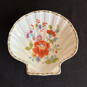 May include: A white, shell-shaped dish with a floral design. The dish features a large orange flower with smaller pink and blue flowers. The rim is decorated with a gold and brown pattern. The dish is approximately 7 inches wide.