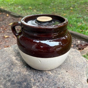 May include: A vintage, two-tone stoneware crock with a lid. The upper portion is a glossy, dark brown, while the lower half is a creamy white. It has a small handle and a round, light-colored lid. The crock sits on a stone surface.