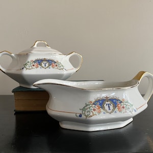 May include: A white porcelain creamer and sugar bowl set with gold trim and floral designs. The bowls feature a blue and gold crest with the letter "T" in the center.