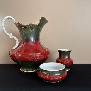 May include: A decorative ceramic pitcher, vase, and bowl set. The pitcher has a white handle and a red, green, and gold color scheme. The vase and bowl share the same color scheme. The set is likely for decorative purposes.