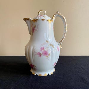 May include: A white porcelain teapot with a floral design in shades of pink and gold. The teapot has a rounded body with a fluted design and a delicate handle.