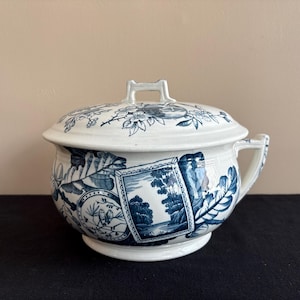 May include: A white and blue ceramic chamber pot with a lid. The pot features a handle and is decorated with blue floral and landscape patterns. The lid has a handle and a floral design. The pot is sitting on a black surface.
