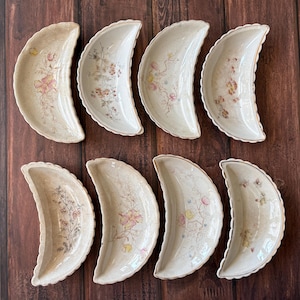 May include: Eight antique, crescent-shaped serving dishes. Each dish features a scalloped edge and a floral pattern in shades of pink, yellow, and green on a cream background. The dishes are arranged on a dark wood surface.