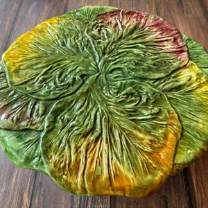 May include: A round ceramic serving dish shaped like a head of lettuce with realistic, textured leaves in shades of vibrant green, yellow, and hints of reddish-purple. The surface features intricate, raised veining and ruffled edges throughout.