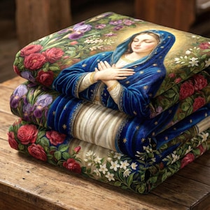 May include: A stack of three decorative blankets featuring a religious image of a woman in a blue robe with a gold halo. The blankets are adorned with floral patterns of roses, irises, and other flowers, with a wooden table in the background.
