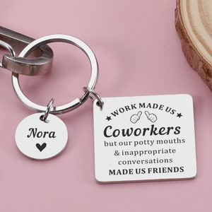 Könnte beinhalten: Silberner Schlüsselanhänger mit einem runden Anhänger, auf dem "Nora" mit einem Herzen steht, und einem quadratischen Anhänger, auf dem steht "Arbeit hat uns zu Kollegen gemacht, aber unsere Schimpfwörter und unsere unangemessenen Gespräche haben uns zu Freunden gemacht".