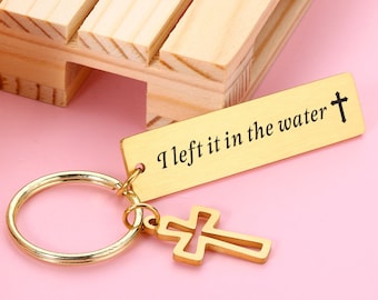 May include: Gold-toned keychain with a cross charm and a rectangular tag that reads "I left it in the water" with a small cross at the end of the text.