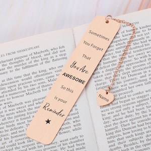 May include: A rose gold bookmark with a quote that reads "Sometimes you forget that you are awesome." The bookmark also has the words "So this is your reminder" and a star. The bookmark is hanging from a chain with a heart-shaped charm that says "Name".