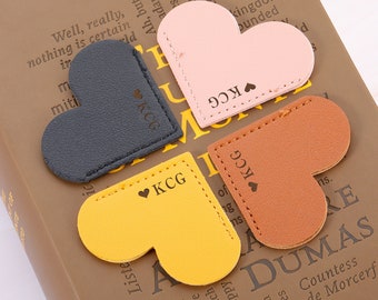 May include: Four heart-shaped leather bookmarks in different colors: black, yellow, pink, and brown. Each bookmark has the letters "KCG" and a small heart symbol engraved on it.