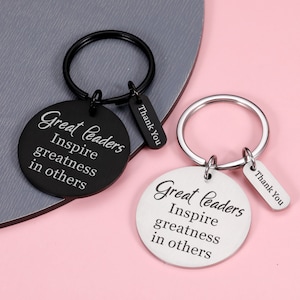 Op de afbeelding: Twee sleutelhangers met ronde metalen labels. De labels zijn zwart en zilver en hebben dezelfde inscriptie: "Great leaders inspire greatness in others." Elke sleutelhanger heeft een klein label met de tekst "Thank You".