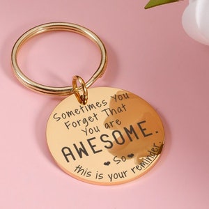 以下が含まれることがあります： ゴールドトーンのキーチェーン。丸いチャームには「Sometimes You Forget That You Are Awesome. So this is your reminder.」と刻まれています。