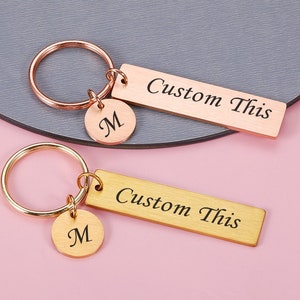 Può includere: Due portachiavi dorati con un ciondolo a barra personalizzato che dice "Custom This" e un piccolo ciondolo rotondo con la lettera "M" incisa.
