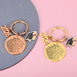 May include: Two circular metal keychains featuring the year 2026 and a graduation cap charm. Each round pendant is engraved with the text: You are Amazing PS. 139:14, Capable Mark 10:27, Chosen 1 Thess. 1:4, Never Alone Matt. 28:20, Always Loved Rom. 8:39, Beautiful Eccl. 3:11, Victorious Rom. 8:37, Enough 2 Cor. 12:9, Created Gen. 1:31, Strong Phil. 4:13, and a cross.