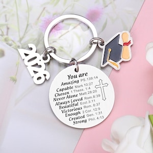 May include: A silver-toned metal keychain featuring a circular pendant engraved with the text You are Amazing PS.139:14, Capable Mark 10:27, Chosen 1 Thess. 1:4, Never Alone Matt.28:20, Always Loved Rom.8:39, Beautiful Eccl.3:11, Victorious Rom. 8:37, Enough 2 Cor. 12:9, Created Gen. 1:31, Strong Phil. 4:13, a cross, a 2026 charm, and a graduation cap charm.