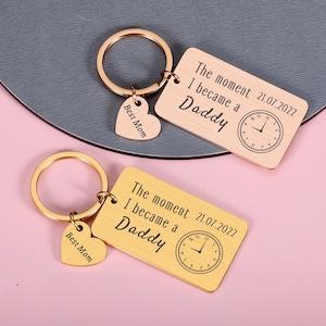 May include: Two rectangular metal keychains with a brushed finish, one rose gold and one gold. Each features a circular ring, a small heart charm engraved with Best Mom, and a plate engraved with The moment I became a Daddy 21.07.2022 and a clock.